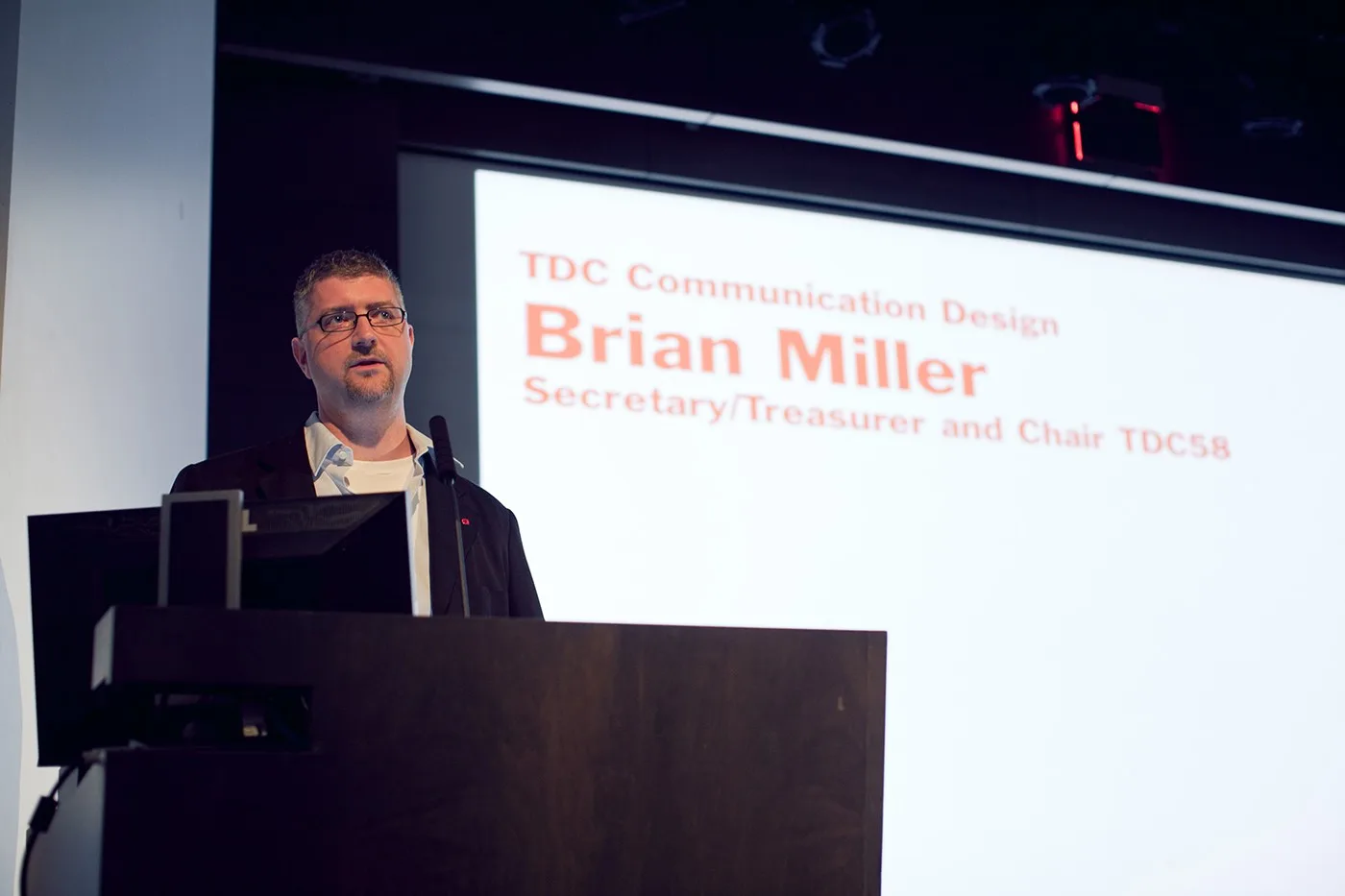 Brian Miller speaking at a podium at the Type Directors Club, in front of a slide reading Secretary/Treasurer and Chair TDC58